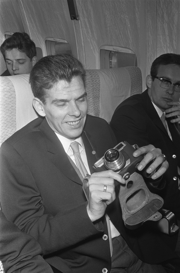 Ein Mann in einem Anzug sitzt in einem Flugzeug und hält eine Kamera in den Händen, flankiert von zwei anderen Männern, von denen einer eine Brille trägt, auf einem Schwarz-Weiß-Bild.