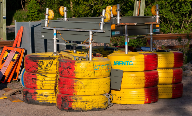 Ein Haufen gelber und roter Reifen auf einem Schmutzfeld umgeben von verstreuten Metallobjekten, Brettern und anderem Schutt, mit Bäumen, Pflanzen und einer Hütte im Hintergrund.