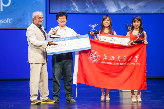 Vier Personen stehen auf einer Bühne, jede hält Auszeichnungen, Schecks und eine Flagge, sie lächeln mit einem digitalen Bildschirm im Hintergrund.