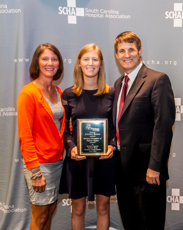 Drei Personen, zwei Frauen und ein Mann, stehen zusammen; der Mann trägt einen schwarzen Anzug, eine Frau trägt ein orange-schwarzes Kleid, und die andere Frau, in einem schwarzen Kleid, hält einen Preis, mit einem grauen Hintergrund.