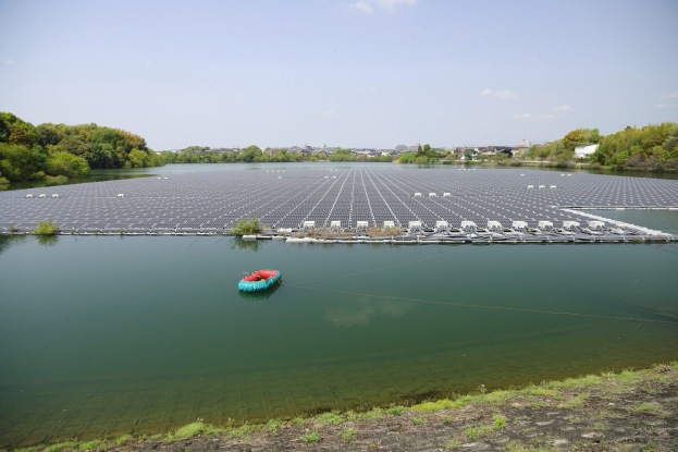 Kleines Boot mit Solarpaneelen auf einem Gewässer, umgeben von Grünflächen, mit Gebäuden und einem klaren blauen Himmel im Hintergrund.