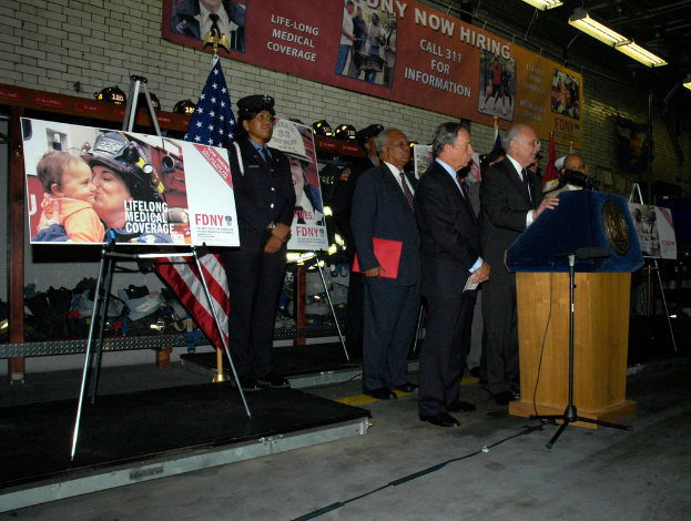 Eine Gruppe von Menschen, die in der Nähe eines Podiums mit Mikrofonen, Schildern auf Ständern, Flaggen und Schildern an der Wand stehen, alles unter hellem Licht.
