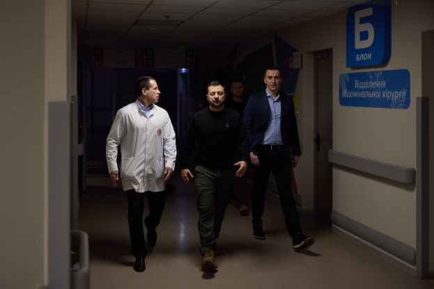 Eine Gruppe von Männern in Not geht einen Krankenhausflur entlang mit Deckenbeleuchtung, vorbei an einer Tür auf der rechten Seite und Texttafeln auf der linken Seite.