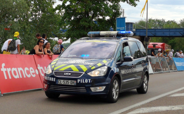 Ein Polizeiwagen fährt auf einer Straße neben einer Menschenmenge, einige tragen Mützen und tragen Taschen, mit einem Banner auf der linken Seite des Wagens und Geländern mit Bannern dahinter; im Hintergrund gibt es Bäume, eine Brücke, eine Fahne auf einem Mast und einen bewölkten Himmel.