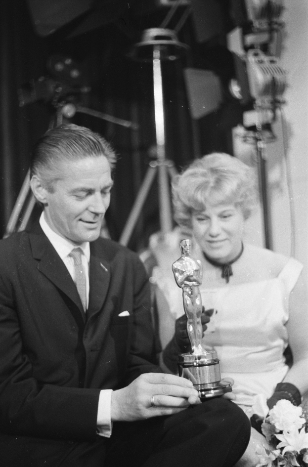 Ein Mann in einem schwarzen Blazer, einem weißen Hemd und einer Krawatte hält eine Oscar-Statue in der Hand, waehrend er neben einer Frau in Abendgarderobe sitzt, die einen Blumenstrauße in der Hand hält, mit weichem Licht im Hintergrund.