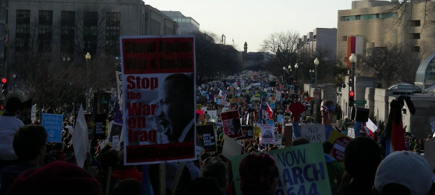 Eine Gruppe von Menschen demonstriert auf einer Stra├če, h├Ąlt Plakate, mit Geb├Ąuden, B├Ąumen, Laternenmasten und dem Himmel im Hintergrund.
