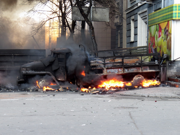 Ein großer Lastwagen brennt auf der Seite einer Straße, wobei dicker Rauch aufsteigt, mit Bäumen, Gebäuden und einem klaren Himmel im Hintergrund.