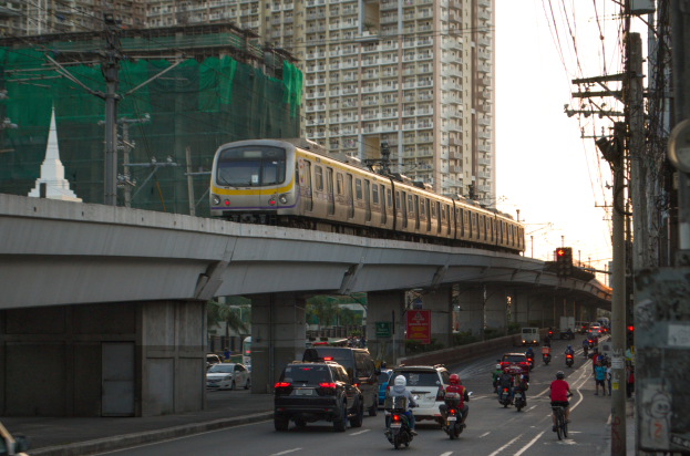 Ein Zug fährt über eine Brücke mit Autos und Motorrädern auf der Straße darunter, Strommasten mit Drähten auf beiden Seiten und Gebäude im Hintergrund.