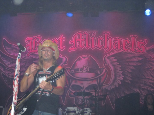 Eine Musik-Konzertszene mit einem Gitarristen, der auf der Bühne spielt, einer Person, die in der unteren rechten Ecke sitzt, einem Banner im Hintergrund und Drogen, die in der Mitte unten platziert sind, mit Bühnenbeleuchtung von oben.