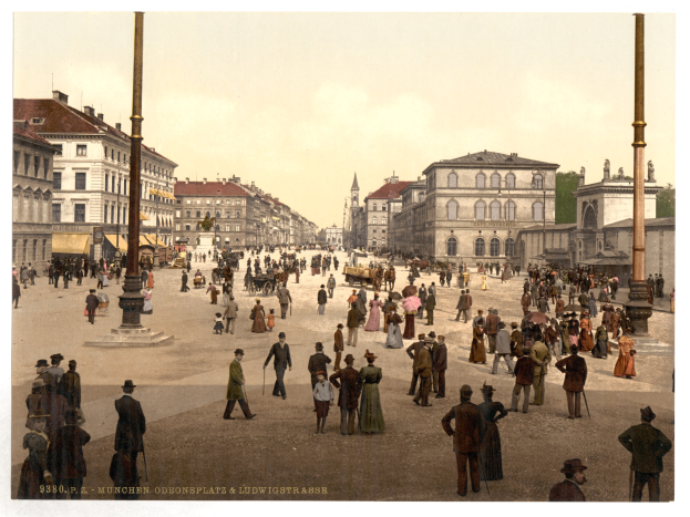 Schwarz-weiß-Foto einer belebten Münchner Straße in den frühen 1900er Jahren mit Fußgängern, einem Pferdewagen, Gebäuden, Bäumen und einem sichtbaren Himmel mit Text am unteren Rand.