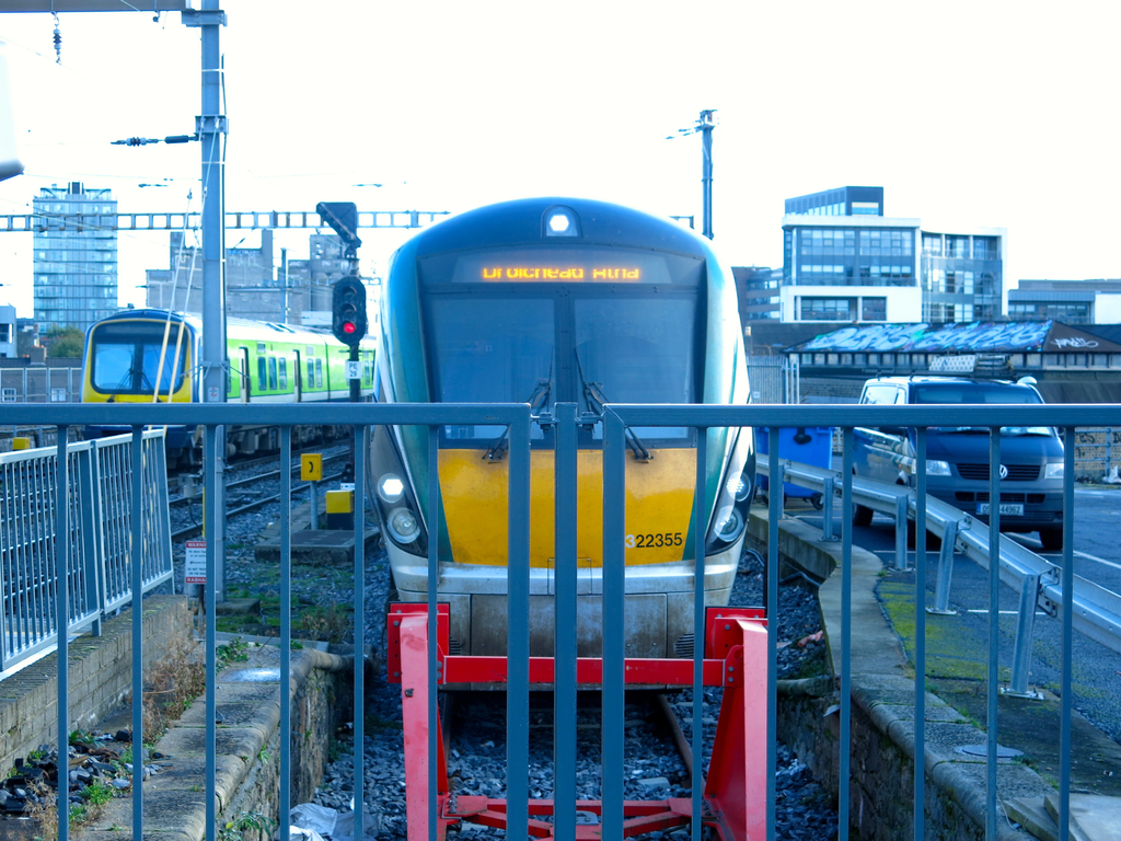 Zwei Züge auf den Schienen mit einem Zaun im Vordergrund, Pfosten mit einem mit einer Signalbeleuchtung, ein Auto auf der Straße rechts, Gebäude im Hintergrund und Himmel oben.