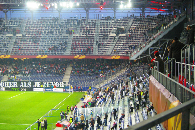 Großes Stadion voller Zuschauer bei einem Fußballspiel, mit sitzenden und stehenden Personen, einer Reling mit einem Banner auf der rechten Seite und einem leeren beleuchteten Stadion im Hintergrund.