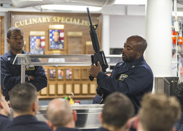 Zwei Polizeibeamte vor einer Gruppe von Menschen stehend, einer hält eine Waffe, mit einer Säule, gerahmten Fotos, Text an einer Wand und Deckenleuchten im Hintergrund.