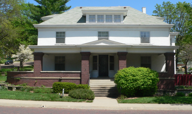 Ein weißes Haus mit einem grünen Dach und einem roten Zaun, umgeben von Pflanzen, Gras, einem Weg, einem Auto auf der Straße, einer Gruppe von Bäumen und einem bewölkten Himmel.