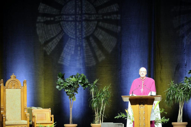 Ein Mann in einem rosa-weißen Kleid steht neben einem Rednerpult in einem Auditorium, mit Pflanzen und zwei Stühlen daneben und einer bunten Wand im Hintergrund.