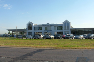 Ein Autohaus mit parkenden Fahrzeugen vorne, umgeben von Gebäuden, Straßenlaternen, fahrenden Fahrzeugen, Infotafeln, Bäumen und einem bewölkten Himmel.