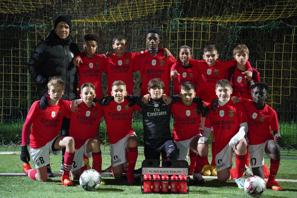 Eine Gruppe junger Jungs in roten T-Shirts und schwarzen Shorts posiert für ein Foto auf einem Fuballfeld, umgeben von Fubällen, einem Netz, einem Zaun und Bäumen im Hintergrund.
