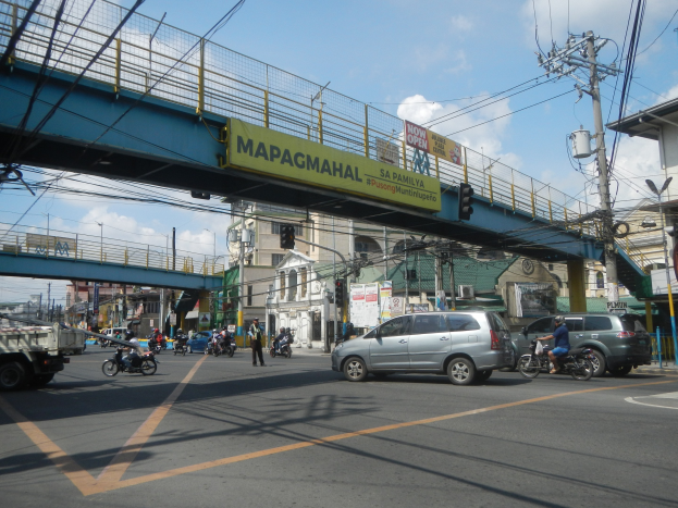 Eine belebte Stadtstraße mit Fahrzeugen, Fußgängern, einer Brücke mit Geländern und einem Namensschild, Strommasten mit Drähten, Laternen, Verkehrsampeln, Gebäuden mit Fenstern und einem bewölkten Himmel.