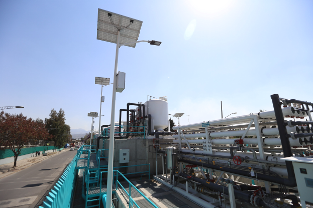 Großes industrielles Gebäude mit ausgedehnten Rohrleitungen und Solarpaneelen auf dem Dach, umgeben von Geländern, Masten, Laternen, Schildern, Bäumen und Fahrzeugen auf der Straße, neben Hügeln unter einem klaren blauen Himmel, Teil der neuen Wasseraufbereitungsanlage von Kalifornien.