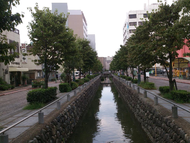 Ein Wasserkanal verläuft durch die Mitte des Bildes, flankiert von Hecken, Bäumen und einem Fußweg auf beiden Seiten, mit Straßen, Gebäuden und fahrenden Autos im Hintergrund unter einem Himmel.