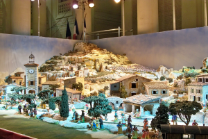 Ein detailliertes Modell einer Krippe, das in einem Museum ausgestellt wird und Figuren, Gebäude, Bäume, eine Uhrenturm, Flaggen und beleuchtete Elemente enthält.