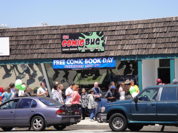 Gruppen von Menschen, die vor einem Gebäude mit Glastüren und einem Dach stehen, zwei Autos auf der Straße, grüne Luftballons, eine Fahne und ein Namensschild auf dem Dach, unter einem klaren Himmel.