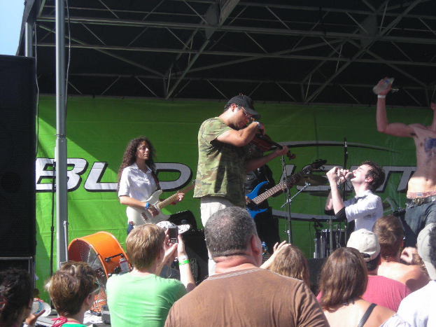 Eine Gruppe von Musikern, die auf der Bühne verschiedene Instrumente spielen, während das Publikum davor steht und die Musik genießt, mit einem großen schwarzen Lautsprecher links und einem grünen Banner im Hintergrund.