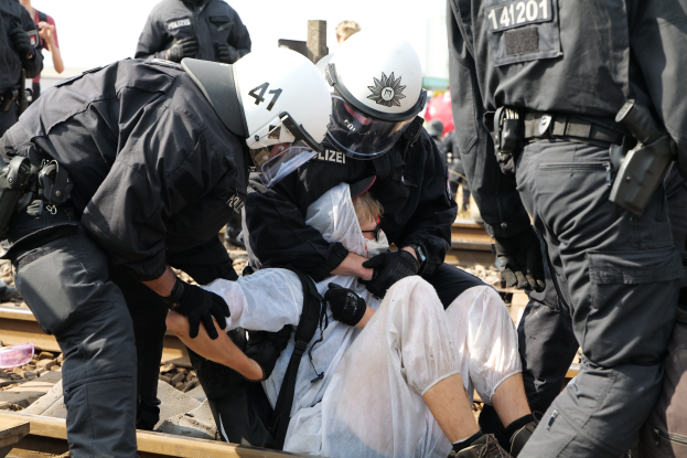 Polizisten in Einsatzausrüstung verhaften einen Mann in einem weißen Kleid auf einem Bahngleis, mit zusätzlichen Beamten und Zuschauern.