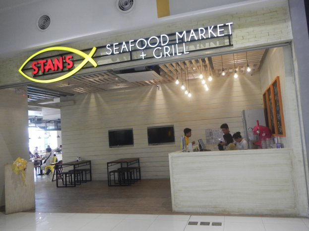 Innenansicht von Stans Seafood Market & Grill mit sitzenden und stehenden Menschen, einem Tisch mit Gegenständen auf der rechten Seite, einem Foto auf der Wand, an der Wand montierten Fernsehern und Deckenbeleuchtung.
