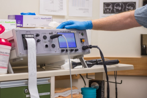 Eine Person in blauen Handschuhen arbeitet an einem medizinischen Gerät auf einem unordentlichen Tisch in einem Raum mit Papieren an der Wand, was auf eine Krankenhausumgebung hinweist.