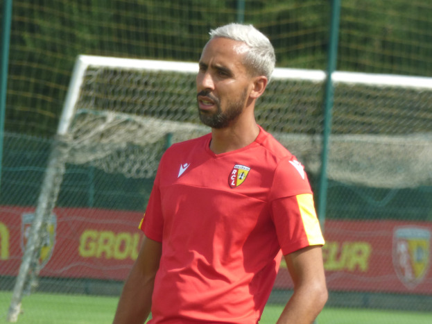 Ein Mann in einem roten T-Shirt steht auf einem Fussballfeld und schaut in die Ferne mit einem Netz und einer "Rajasthan FC"-Fahne im Hintergrund.