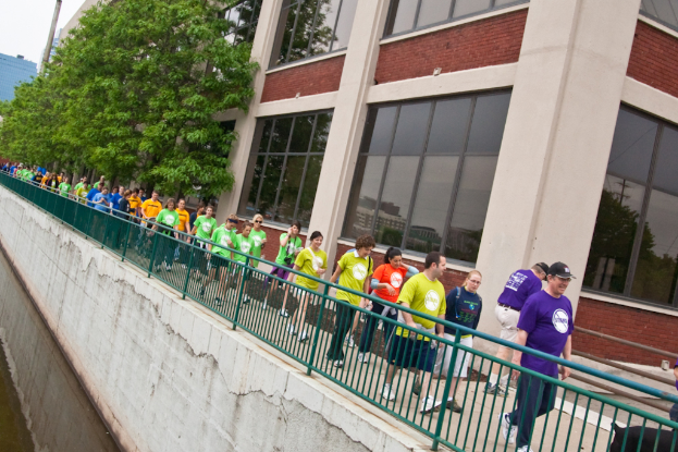 Einige Menschen gehen auf der rechten Seite eines Weges entlang einer Reling, mit einem Gebäude mit Glasfenstern und Bäumen im Hintergrund, einem klaren Himmel darüber und einem See auf der linken Seite.