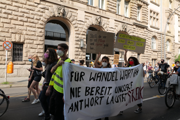 Eine Gruppe von Menschen marschiert auf der Straße in Berlin, hält Schilder und Banner hoch und fährt Fahrräder, vor einem Gebäude mit Fenstern, Bögen, Säulen, Skulpturen, Bäumen und einem Laternenpfahl, an einer Anti-Kriegs-Demonstration teilnehmend.