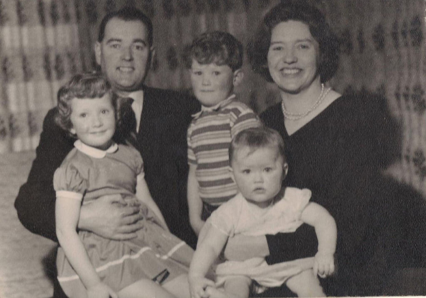 Eine Familie mit fünf Personen, einem Mann in Anzug und Krawatte links, einer Frau rechts und drei strahlenden Kindern in der Mitte.