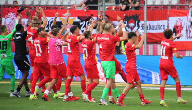 Eine Gruppe von Fußballspielern feiert ein Tor auf einem Feld, einige halten Flaschen und andere klatschen, mit einem Zaun, Bannern, Metallstangen, Stühlen und einer Decke mit Deckenleuchten im Hintergrund.