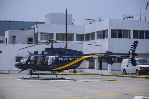 Ein Polizei-Hubschrauber vor einem Gebäude geparkt, daneben ein Van, Strommasten und Leitungen im Hintergrund und der Himmel oben.