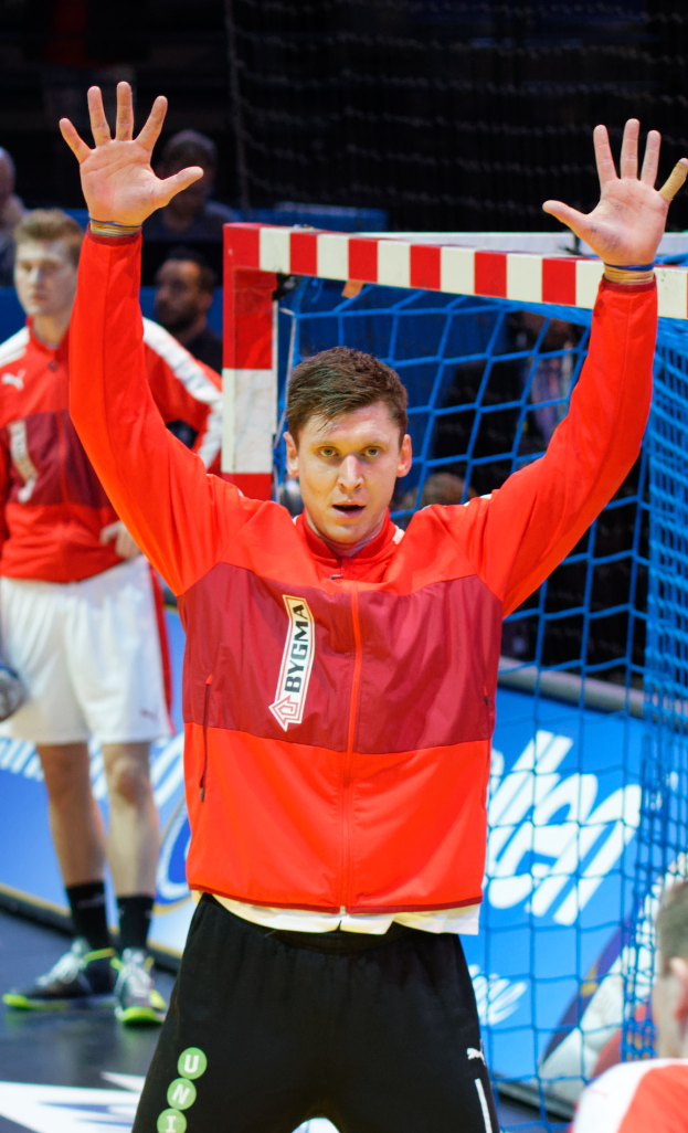 Ein Mann in einer roten Jacke und schwarzen Shorts steht auf einem Futsal-Platz, trägt Schuhe und hat ein Netz sowie einige Personen im Hintergrund.
