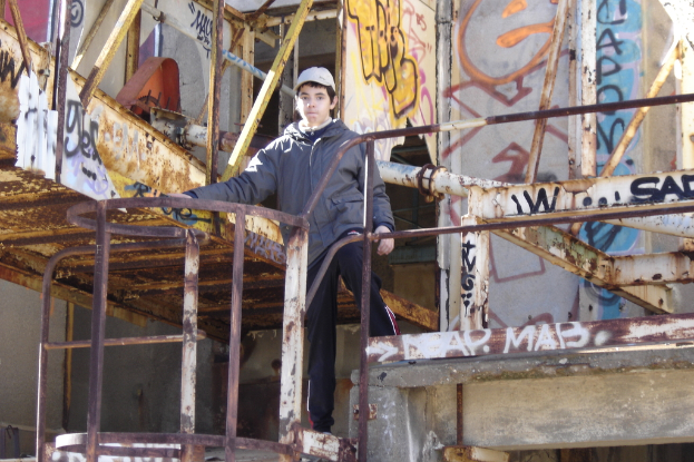 Ein Junge in einer Jacke und einer Mütze steht vor einem Metallblockzaun mit Graffiti an der Wand.