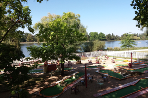 Minigolfanlage in einem Park mit grünem Gras, Bäumen, einem Zaun, verstreuten Stühlen und Tischen, einem Gewässer mit einem Boot im Hintergrund und sichtbarem Himmel.