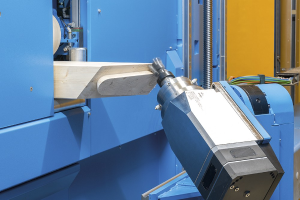 CNC-Fräsmaschine schneidet ein Holzstück in einer industriellen Umgebung mit sichtbaren Kabeln und einer Wand im Hintergrund.