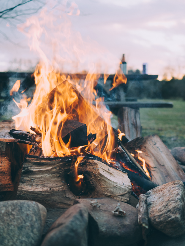 Ein Lagerfeuer brennt hell in einem Feld, umgeben von Holzscheiten und Steinen, mit einer Bank, Bäumen und einem klaren blauen Himmel im Hintergrund.