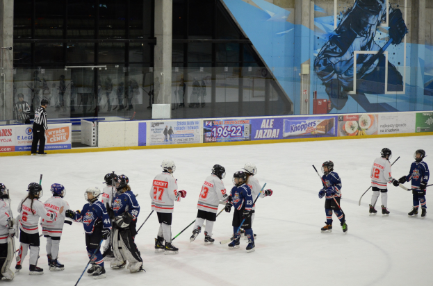 Gruppe junger Menschen beim Eishockey auf einer Indoor-Eisbahn, mit Helmen und Stöcken, einer Person links daneben und Bannern, Säulen und Glaswänden im Hintergrund.
