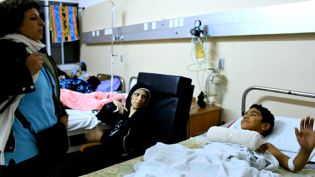 Ein Krankenhauszimmer mit besetzten Betten, Infusionsflaschen auf Ständern zwischen den Betten und einer Frau mit einer Tasche in der Nähe eines Patienten.