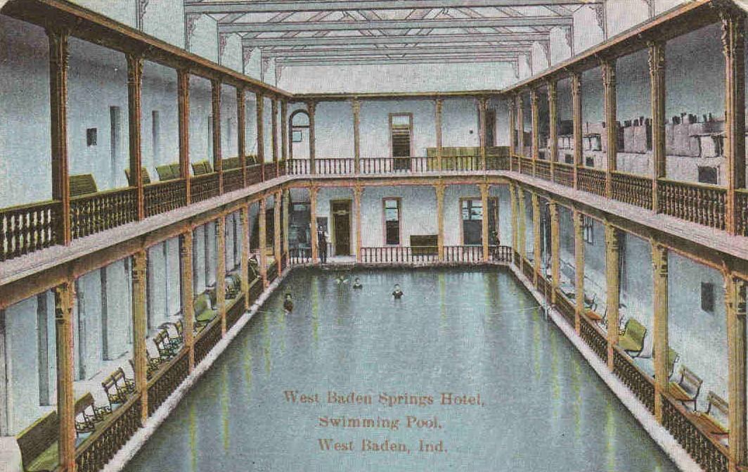 Schwarz-weiß-Postkarte des West Baden Springs Hotels mit einem großen Indoor-Schwimmbad und Menschen im Wasser, umgeben von architektonischen Details und Text am unteren Rand.