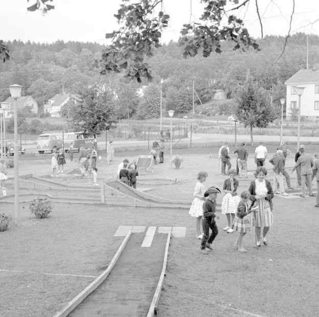 Schwarzes und weißes Bild von Menschen, die Minigolf in einem Park mit Grünflächen, Laternen, geparkten Fahrzeugen und Gebäuden im Hintergrund spielen.