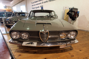 Ein klassischer Alfa Romeo Giulietta Sprint GT Junior aus den 1970er Jahren, der in einem Museum ausgestellt ist und von abgesperrten Pfosten umgeben ist, mit warmer Deckenbeleuchtung und Text an der Hintergrundwand.