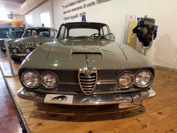 Ein klassischer Alfa Romeo Giulietta Sprint GT Junior aus den 1970er Jahren, der in einem Museum ausgestellt ist und von abgesperrten Pfosten umgeben ist, mit warmer Deckenbeleuchtung und Text an der Hintergrundwand.