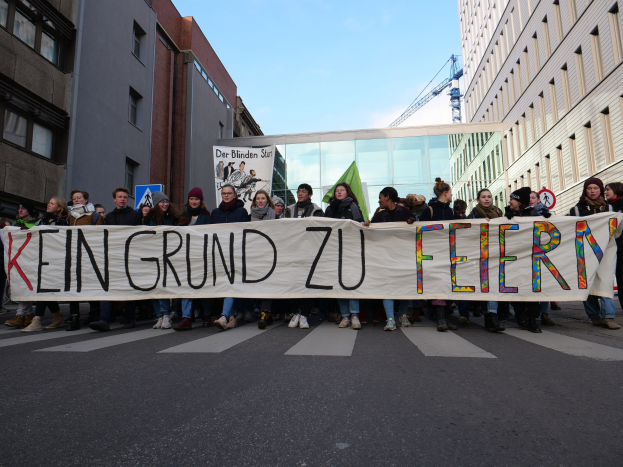 Demonstranten auf einer Straße mit einem Banner "Kein Grund zu Feiern" gegen deutsche Sparmaßnahmen, mit Gebäuden, einem Kran und einem bewölkten Himmel im Hintergrund.