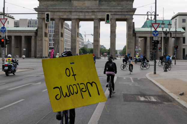 Eine Gruppe von Menschen in Helmen fährt mit Fahrrädern vor dem Brandenburger Tor in Berlin vorbei, während im Hintergrund Laternenpfähle, Verkehrsampeln, Gebäude, Bäume und ein klarer blauer Himmel zu sehen sind.