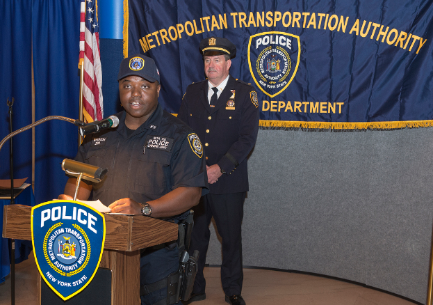 Zwei Polizeibeamte stehen vor einem Rednerpult, einer hält Papiere und spricht in ein Mikrofon, daneben eine Fahne, ein Tisch und eine Tafel mit Symbolen und Text im Hintergrund.
